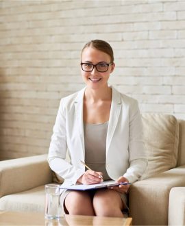 A woman in a white blazer and glasses sits on a cream-colored sofa in a well-lit room, exuding professionalism and warmth. With a pen and clipboard ready, she offers online Suboxone treatment guidance. A wooden shelf with baskets adds charm to the serene setting.