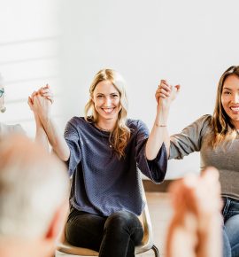 In a bright room in Fort Wayne, a diverse group sits in chairs, holding hands and smiling. The focus is on three individuals—two women and one older woman with white hair—enjoying a moment of unity and warmth, perhaps discussing online detox or pain management strategies.