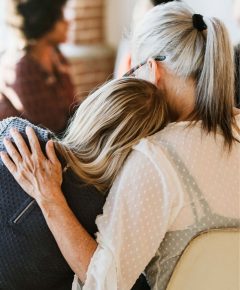 A person with gray hair pulled back in a ponytail embraces a younger individual who has blonde hair, resting their head on the older person's shoulder. They sit closely together in a softly lit room, discussing pain management while others are present in the background.