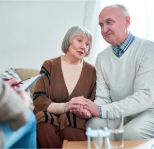 An elderly couple sits on a sofa in Fort Wayne, holding hands. They appear engaged in a serious conversation about online detox options. A person with a clipboard sits opposite them, partially visible. Glasses of water are on a table in the foreground.