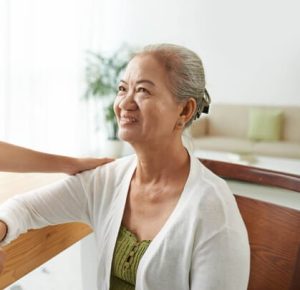 An elderly woman with gray hair, wearing a white cardigan and green top, smiles while sitting at a table. In the warmth of her Fort Wayne home, she receives a comforting hand on her shoulder. A plant and a sofa with green pillows are in the background, creating an atmosphere of solace and peace.